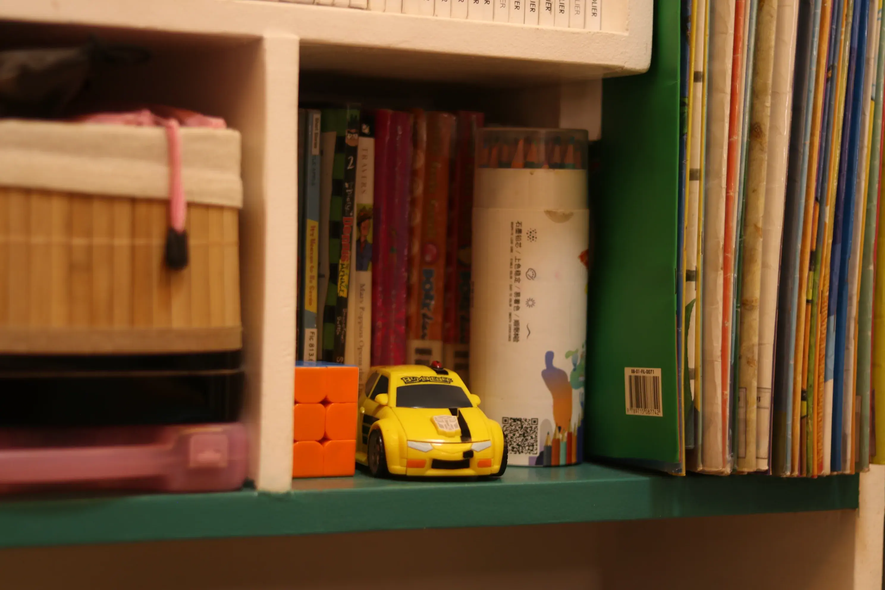 A green shelf with children's books, colored pencils, a puzzle cube, and a yellow toy car