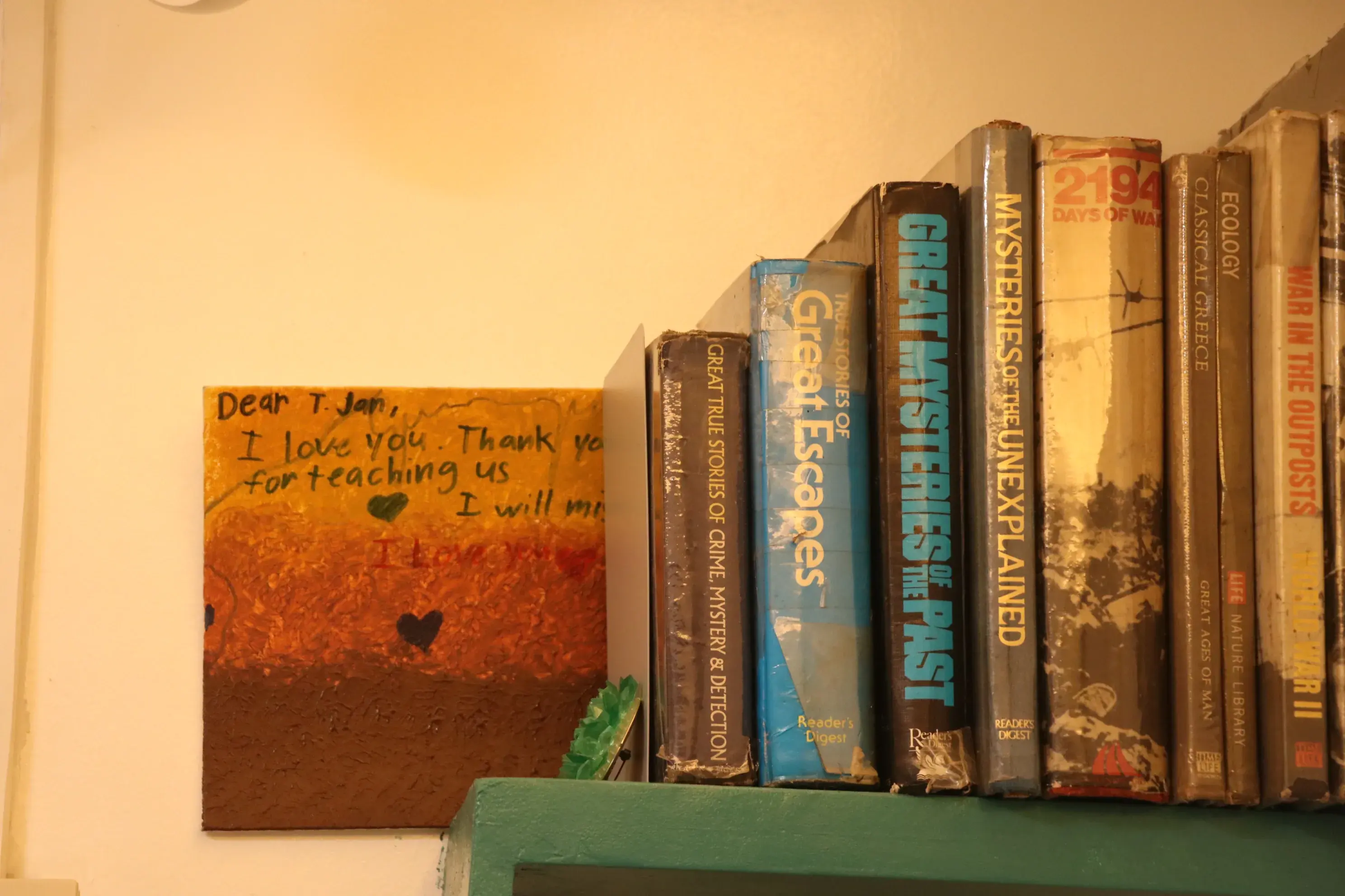 A student thank-you painting next to worn books on a green shelf